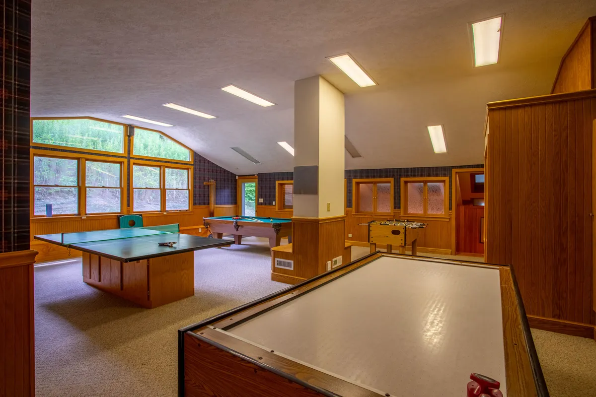 This image showcases a lively game room filled with a pool table, ping pong table, and foosball table, all set against a backdrop of large windows. The space appears open and bright, inviting group play and social gatherings.