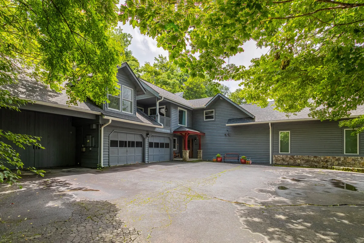 The photo showcases the entrance to the home with its striking red door, attached garage, and surrounding trees. The driveway area suggests a gathering place for friends and family, with mature trees providing dappled shade and a sense of privacy. The lush greenery hints at pleasant spring and summer days spent outdoors.