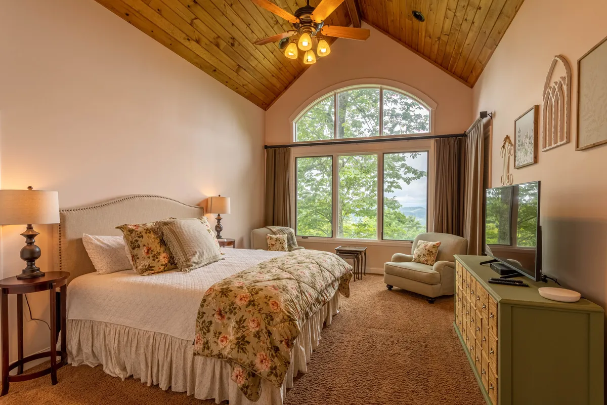 A serene bedroom with a large, comfortable bed positioned to face expansive windows that let in natural light and offer glimpses of the outdoors. A decorative armchair sits in the corner, and a TV console completes the restful scene. The soft color palette and uncluttered design emphasize relaxation and comfort.