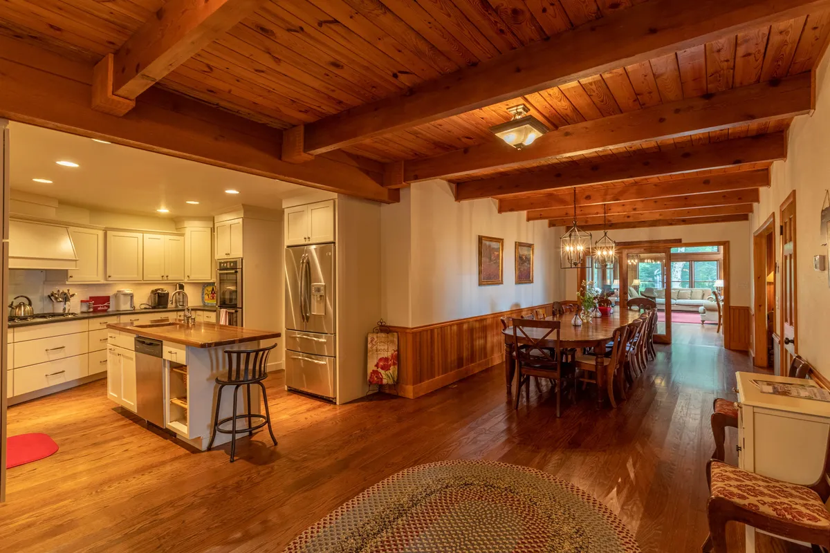 This image showcases a spacious kitchen and dining area featuring a prominent kitchen island, a large dining table that can accommodate many guests, exposed wooden beams, and expansive windows letting in natural light. The scene suggests an open, sociable environment perfect for group gatherings and celebrations.