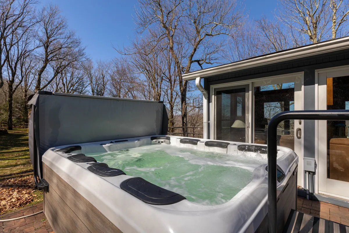 An exterior view of the deck showcasing a bubbling hot tub, surrounded by tranquil trees. The deck offers space for socializing or stargazing, with easy access from the interior living spaces.