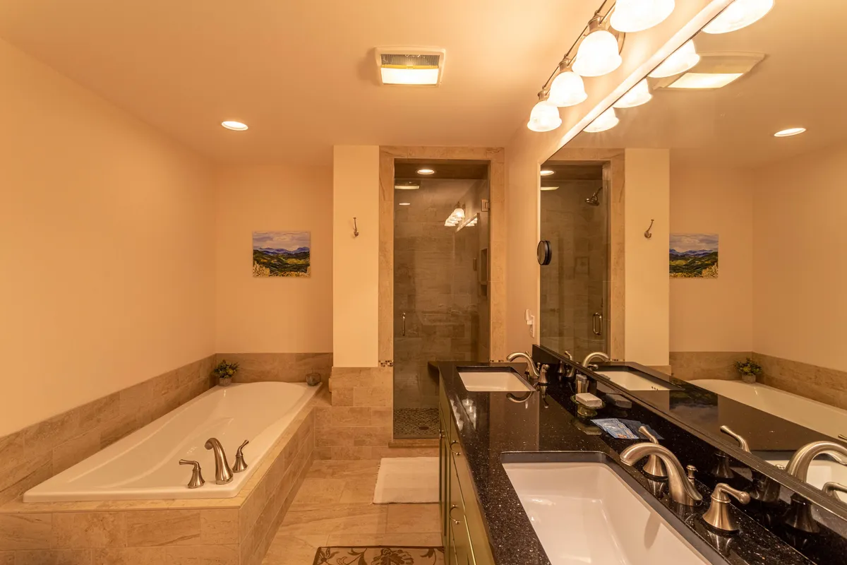 A spacious bathroom featuring a freestanding soaking tub, a double sink granite vanity, a glass walk-in shower, and a landscape painting. The space is awash with natural light, exuding tranquility and spa-like luxury.