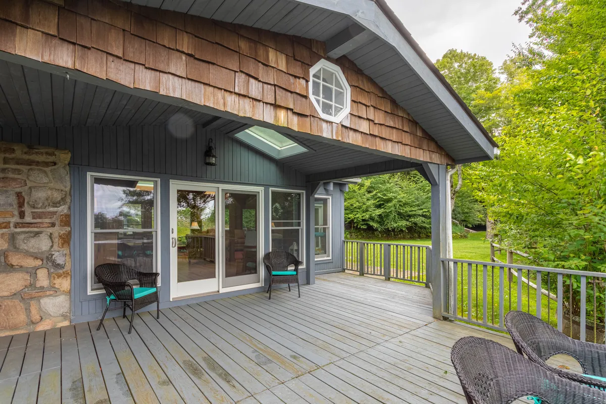 The photo features a wooden deck with large sliding glass doors, a shingle roof, and lush greenery in the background. The deck offers plenty of space for socializing and easily connects the home's interior to its natural surroundings.