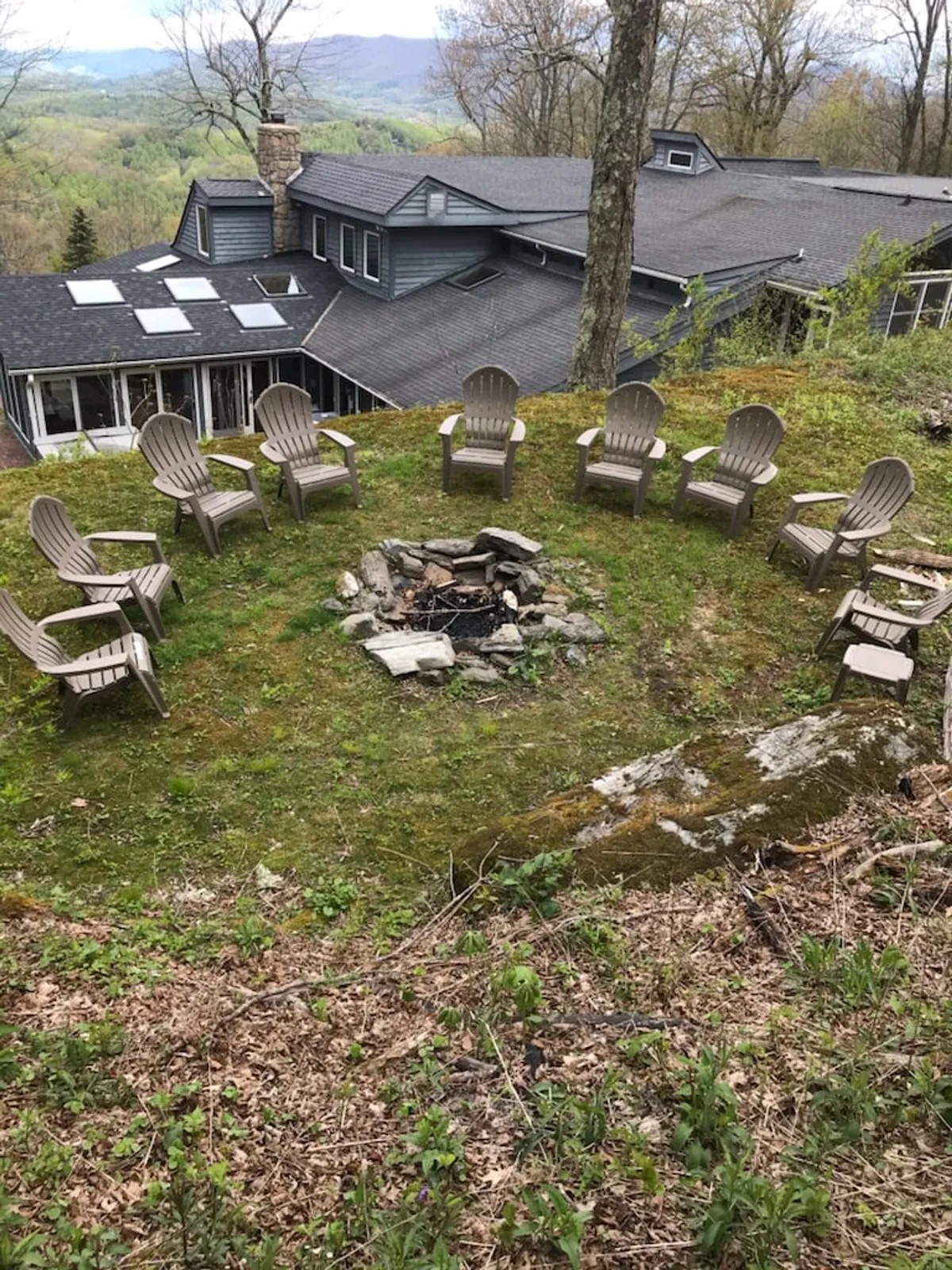 A cozy fire pit encircled by Adirondack chairs sits against a backdrop of sweeping mountain views and natural landscape. The setting exudes warmth and relaxation, inviting guests to gather for campfire stories, marshmallow toasting, and stargazing after a day of exploration.