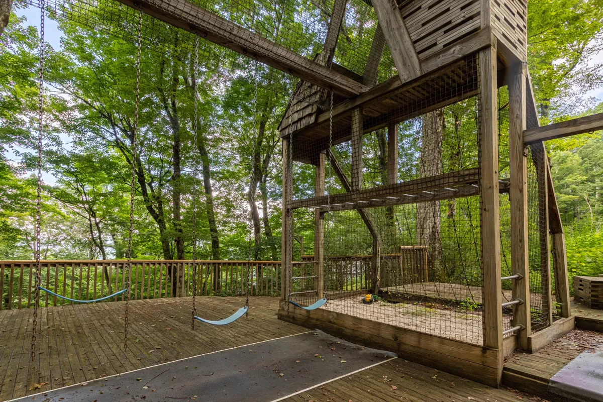 A spacious play structure stands nestled among tall trees, featuring swings, a wooden deck, and ample space for children to run and play. The surrounding forested landscape creates a sense of adventure and privacy, while the sturdy construction invites children and families to spend time outdoors.