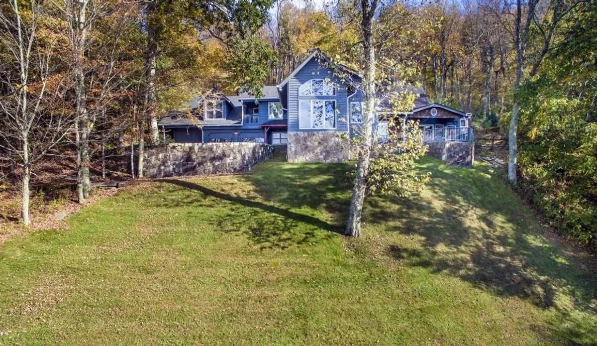 The hero image features a grand, mountain-style single-family home perched amid lush Appalachian trees. A sloped green lawn stretches toward the house, which boasts a spacious wraparound porch perfect for taking in sweeping sunset views. The scene exudes tranquility, with natural wood accents and an inviting, peaceful atmosphere that welcomes groups and families to gather outdoors.